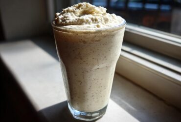 A tall glass filled with a speckled Homemade Vanilla Bean Frappuccino topped with whipped cream, backlit by a window.