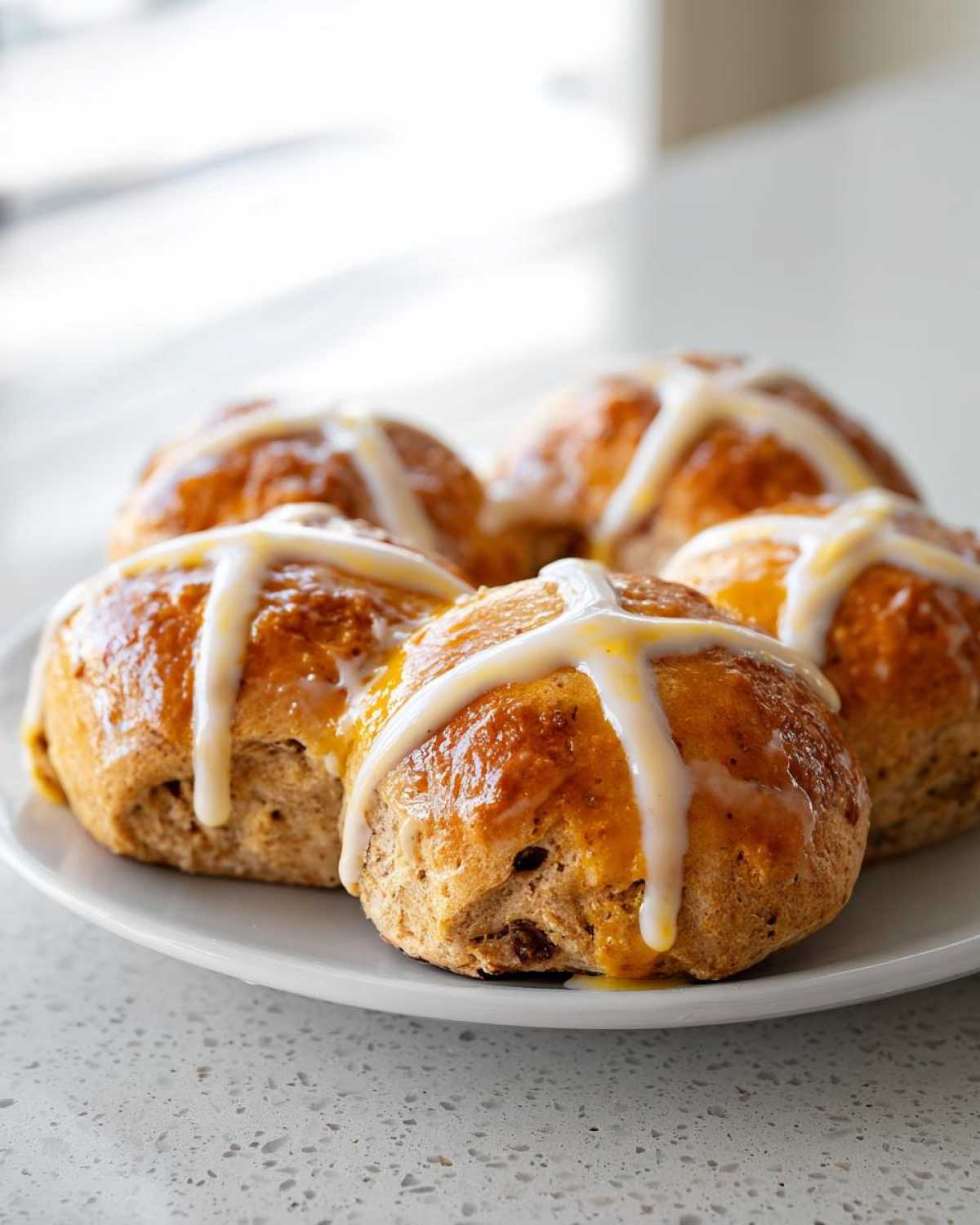 A close-up of four freshly baked Hot Cross Buns with Orange Glaze drizzled across the top, resting on a white plate.