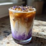 A tall glass of Iced Lavender Latte showing a beautiful purple swirl at the bottom, topped with ice and fresh lavender buds.