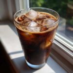 A tall glass filled with a dark, cold Iced Mocha and large ice cubes, resting on a sunny windowsill.