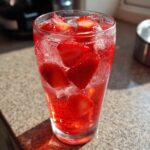 Close-up of a tall glass filled with vibrant red Iced Strawberry Lemon Tea, ice, and fresh strawberry slices.