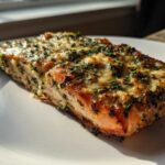 A perfectly cooked fillet of Keto Garlic Butter Salmon topped with melted cheese and herbs, resting on a white plate.