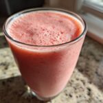 A close-up of a pink, frothy Keto Strawberry Cheesecake Smoothie served in a clear glass on a granite countertop.