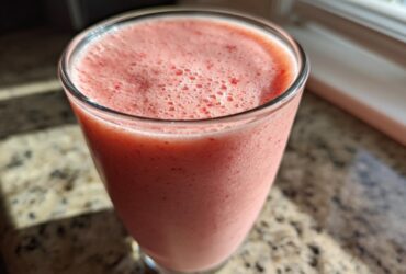 A close-up of a pink, frothy Keto Strawberry Cheesecake Smoothie served in a clear glass on a granite countertop.