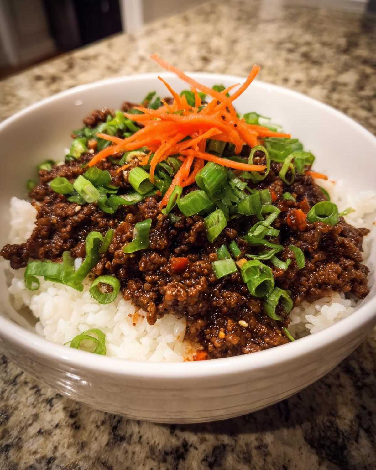 Close-up of a white bowl filled with white rice topped with rich Korean Ground Beef Bulgogi and garnished with green onions and shredded carrots.