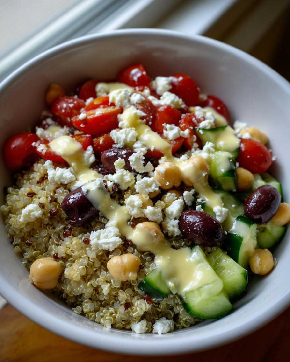 Close-up of a Mediterranean Quinoa Bowl topped with cherry tomatoes, feta, cucumbers, olives, and lemon tahini sauce.