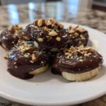 A stack of No-Bake Chocolate Covered Banana Bites dipped in dark chocolate and topped with chopped nuts on a white plate.
