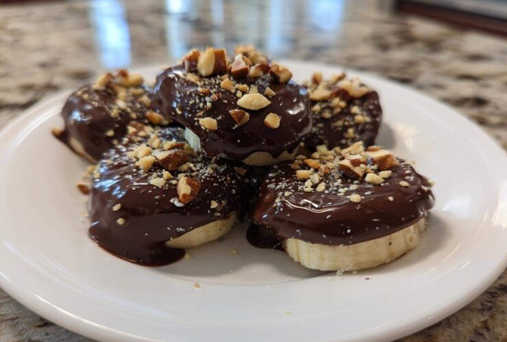 A stack of No-Bake Chocolate Covered Banana Bites dipped in dark chocolate and topped with chopped nuts on a white plate.