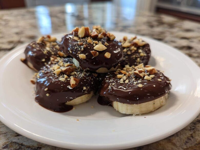 A stack of No-Bake Chocolate Covered Banana Bites dipped in dark chocolate and topped with chopped nuts on a white plate.