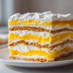 A tall slice of No-Bake Lemon Icebox Cake showing layers of graham crackers, bright yellow lemon filling, and white whipped cream.