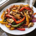 A white plate filled with colorful strips of seasoned chicken, red, yellow, green peppers, and purple onions from One-Pan Chicken Fajitas with Peppers and Onions.