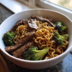 Close-up of a white bowl filled with steaming One-Pot Beef and Broccoli Ramen Noodles, featuring savory beef strips and bright green broccoli florets.