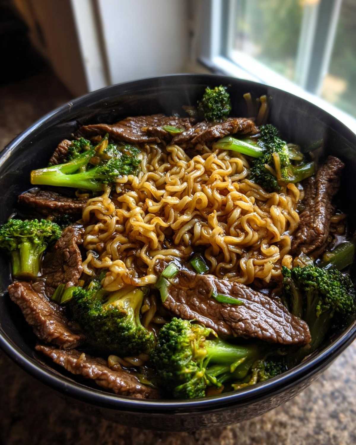 Close-up of a steaming black bowl filled with One-Pot Beef and Broccoli Ramen Noodles, featuring curly noodles, sliced beef, and bright green broccoli florets.