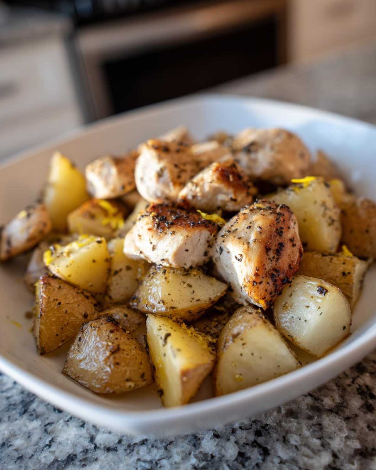 Close-up of seasoned chicken pieces and roasted potatoes served in a white bowl, featuring lemon pepper seasoning.