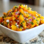 Close-up of a white bowl filled with bright orange Spicy Mango Salsa, featuring diced mango, red onion, and cilantro.