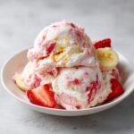 Three scoops of marbled pink and white Strawberry Banana Ice Cream garnished with fresh strawberry slices and banana coins.