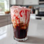 A tall glass filled with layers of dark cold brew coffee, creamy foam, and bright red strawberry syrup drizzle, making a Strawberry Cream Cold Brew.