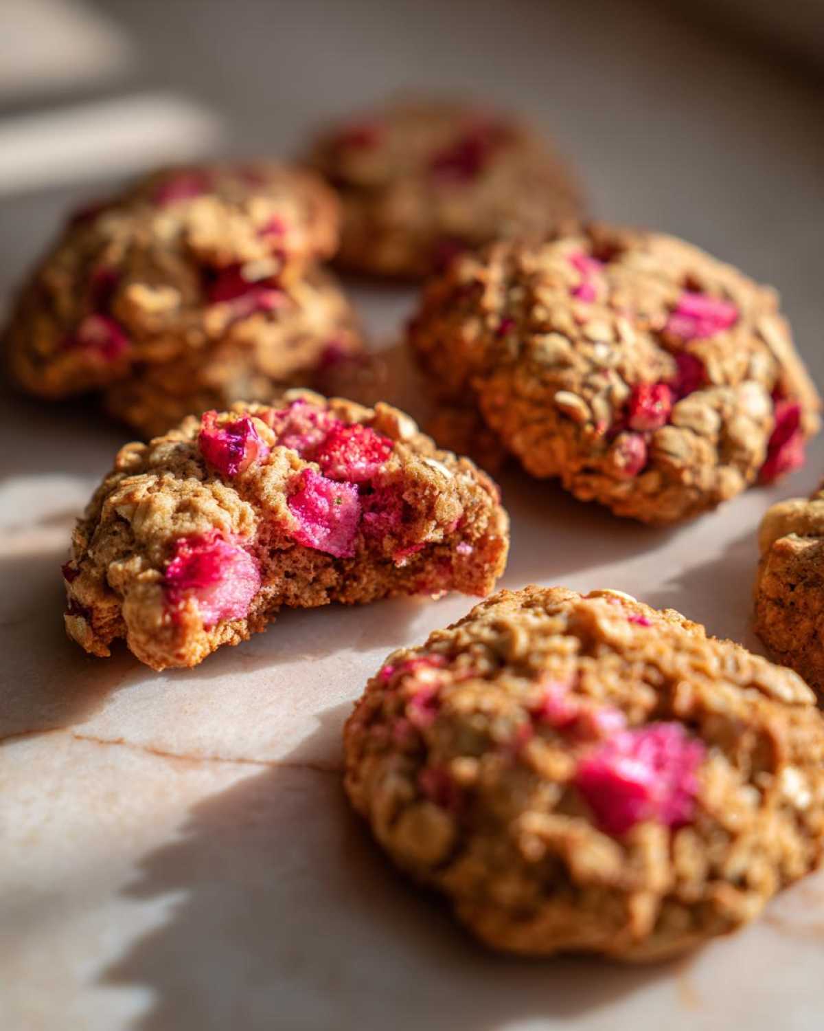 Several golden brown Strawberry Oatmeal Breakfast Cookies studded with bright pink strawberry pieces on a marble surface.