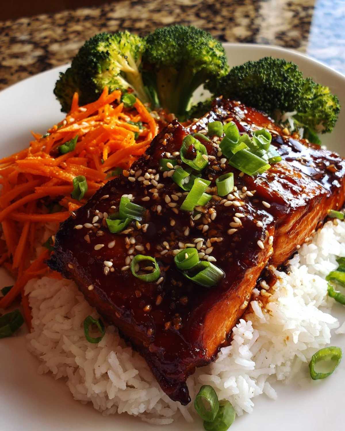 A close-up of a Teriyaki Salmon Rice Bowls serving featuring glazed salmon over white rice, shredded carrots, and broccoli.