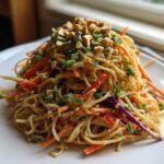 A mound of Thai Peanut Noodles with Crunchy Veggies, topped with chopped peanuts and green onions, served on a white plate.