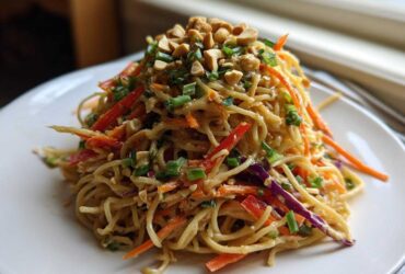A mound of Thai Peanut Noodles with Crunchy Veggies, topped with chopped peanuts and green onions, served on a white plate.
