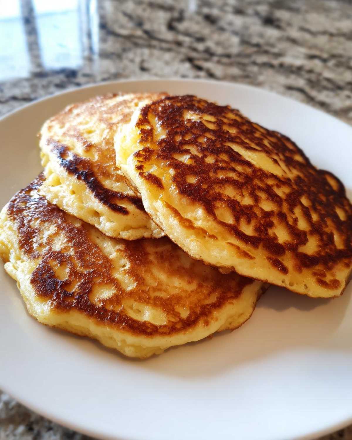 Three golden-brown Vegan Banana Oat Pancakes stacked on a white plate, showing a perfectly cooked, slightly textured surface.