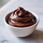 A close-up of rich, dark Vegan Chocolate Avocado Mousse swirled high in a small white bowl on a marble surface.