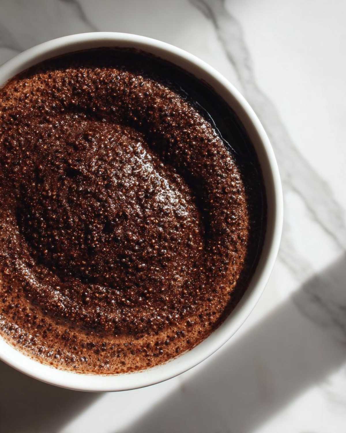 Overhead view of rich, dark Vegan Chocolate Chia Pudding with a bubbly texture in a white bowl on marble.
