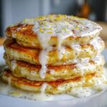 A tall stack of fluffy Vegan Lemon Poppy Seed Pancakes drizzled with thick white glaze and lemon zest.