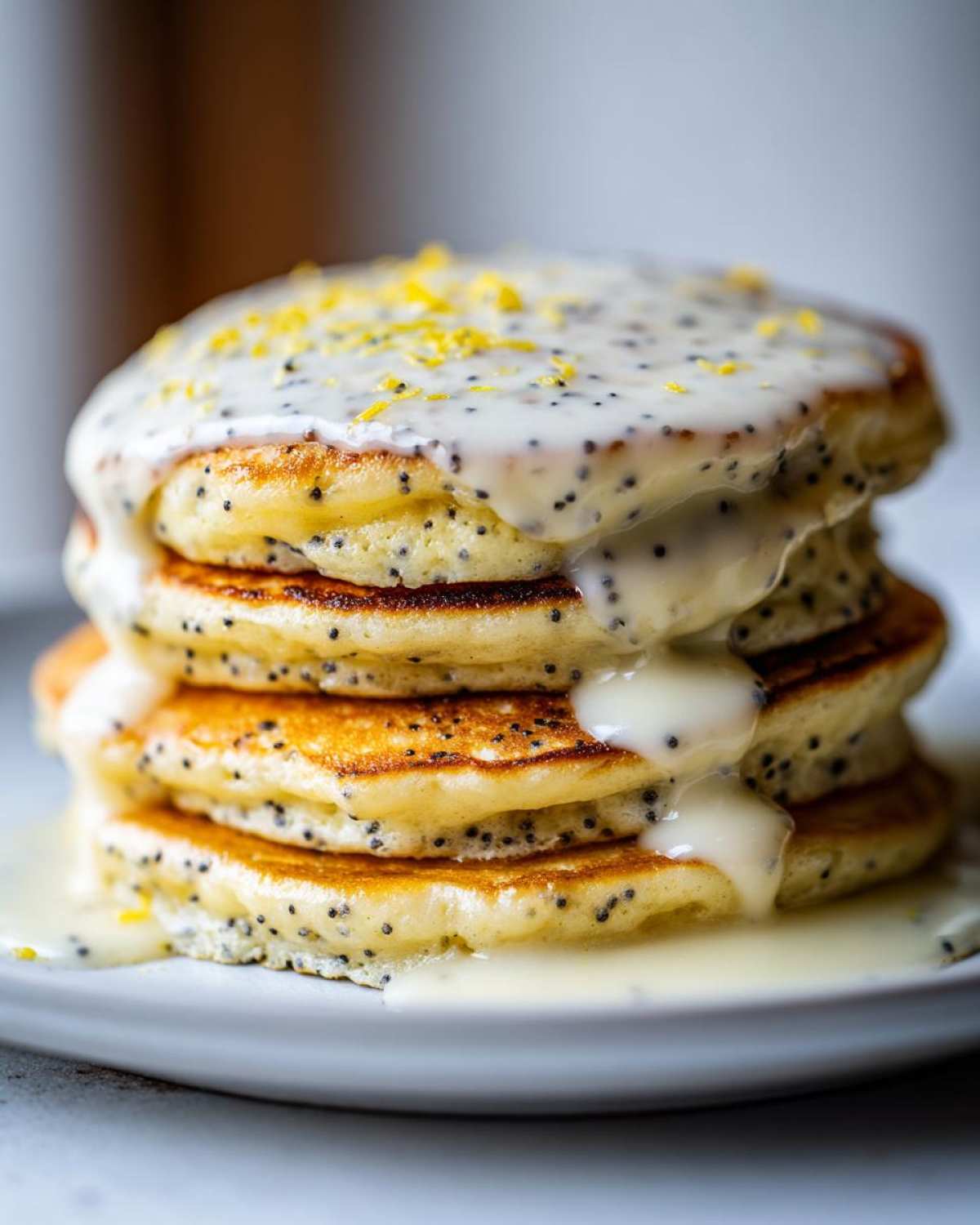 A tall stack of fluffy Vegan Lemon Poppy Seed Pancakes drizzled with lemon glaze and zest.