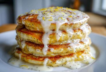 A tall stack of fluffy Vegan Lemon Poppy Seed Pancakes drizzled with thick white glaze and lemon zest.