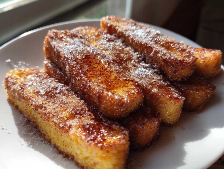 Close-up of golden Air Fryer French Toast Sticks coated heavily in sparkling cinnamon sugar on a white plate.