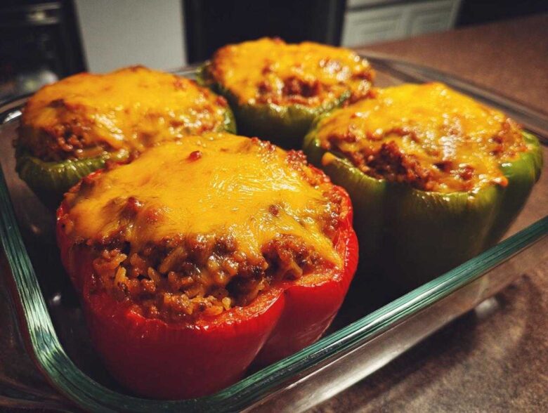 Four Cheesy Ground Beef Stuffed Peppers with Rice, topped with melted cheddar cheese, resting in a glass baking dish.