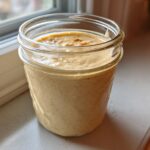 A glass mason jar filled with creamy, beige Copycat Olive Garden Salad Dressing, sitting on a windowsill.