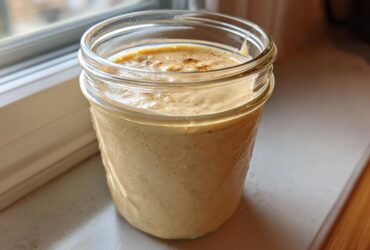 A glass mason jar filled with creamy, beige Copycat Olive Garden Salad Dressing, sitting on a windowsill.