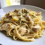 A close-up of fettuccine coated in rich sauce, topped with seasoned chicken chunks, in Creamy Alfredo Pasta with Chicken and Cheese.