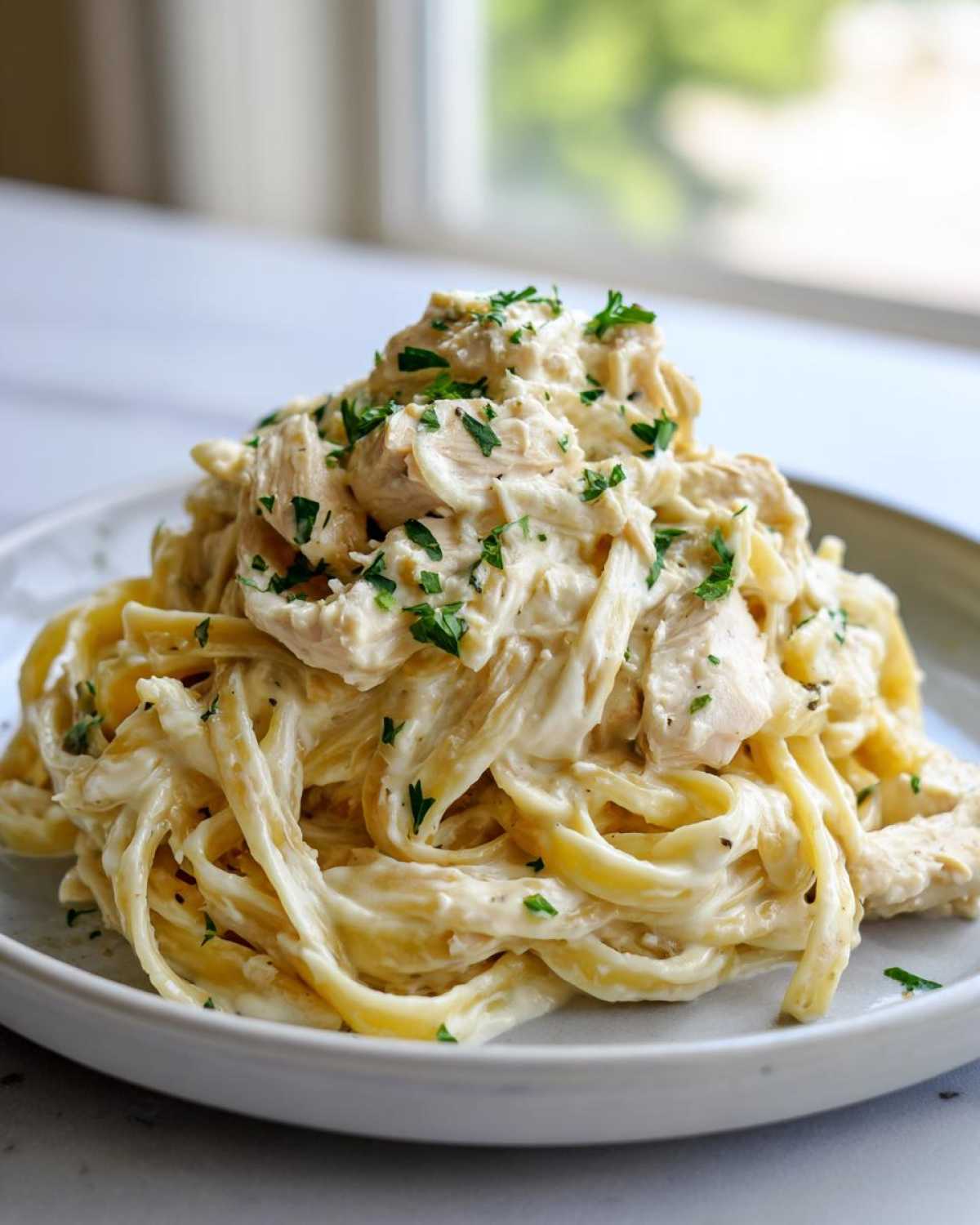 A mound of fettuccine noodles coated in rich, creamy sauce, topped with chunks of chicken and fresh parsley for Creamy Alfredo Pasta with Chicken and Cheese.