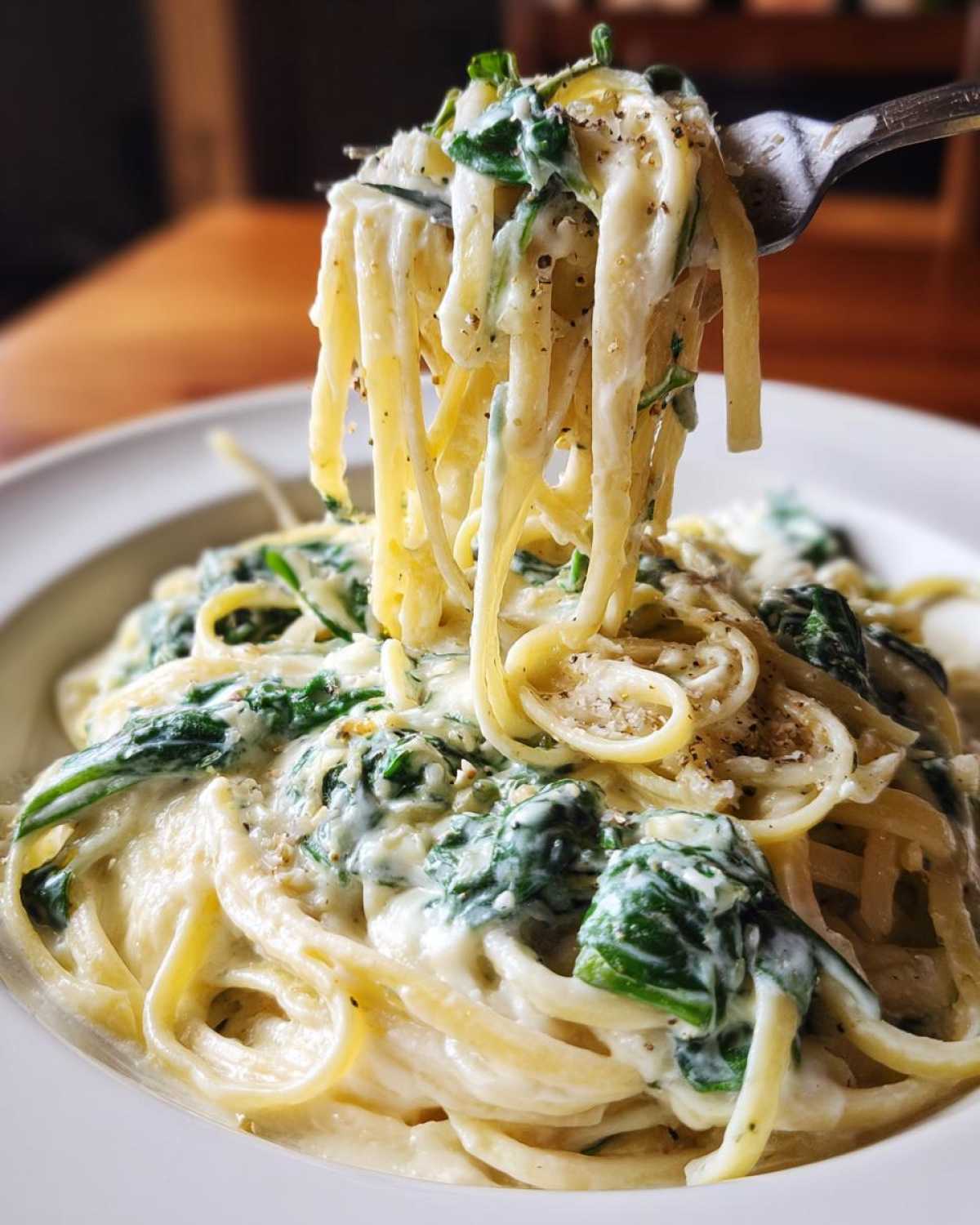 Close-up of a fork lifting creamy linguine pasta mixed with wilted spinach, seasoned with black pepper, from a bowl of Creamy Lemon Ricotta Pasta with Spinach.