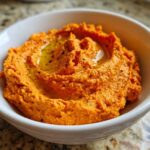 Close-up of vibrant orange Roasted Red Pepper Hummus, swirled and drizzled with olive oil, served in a white bowl.
