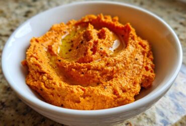 Close-up of vibrant orange Roasted Red Pepper Hummus, swirled and drizzled with olive oil, served in a white bowl.