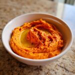 A close-up of creamy Roasted Red Pepper Hummus swirled in a white bowl, topped with olive oil and paprika.