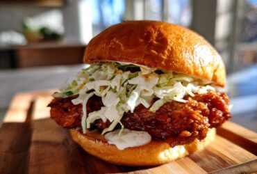 Close-up of a Crispy Chicken Sandwich with Creamy Slaw piled high on a toasted brioche bun, resting on a wooden board.