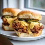 Two Crispy Chicken Sliders topped with melted cheese sauce and pickles, ready to eat.