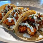Three Crispy Chicken Tacos with Garlic Lime Sauce drizzled over breaded chicken pieces and topped with cilantro.