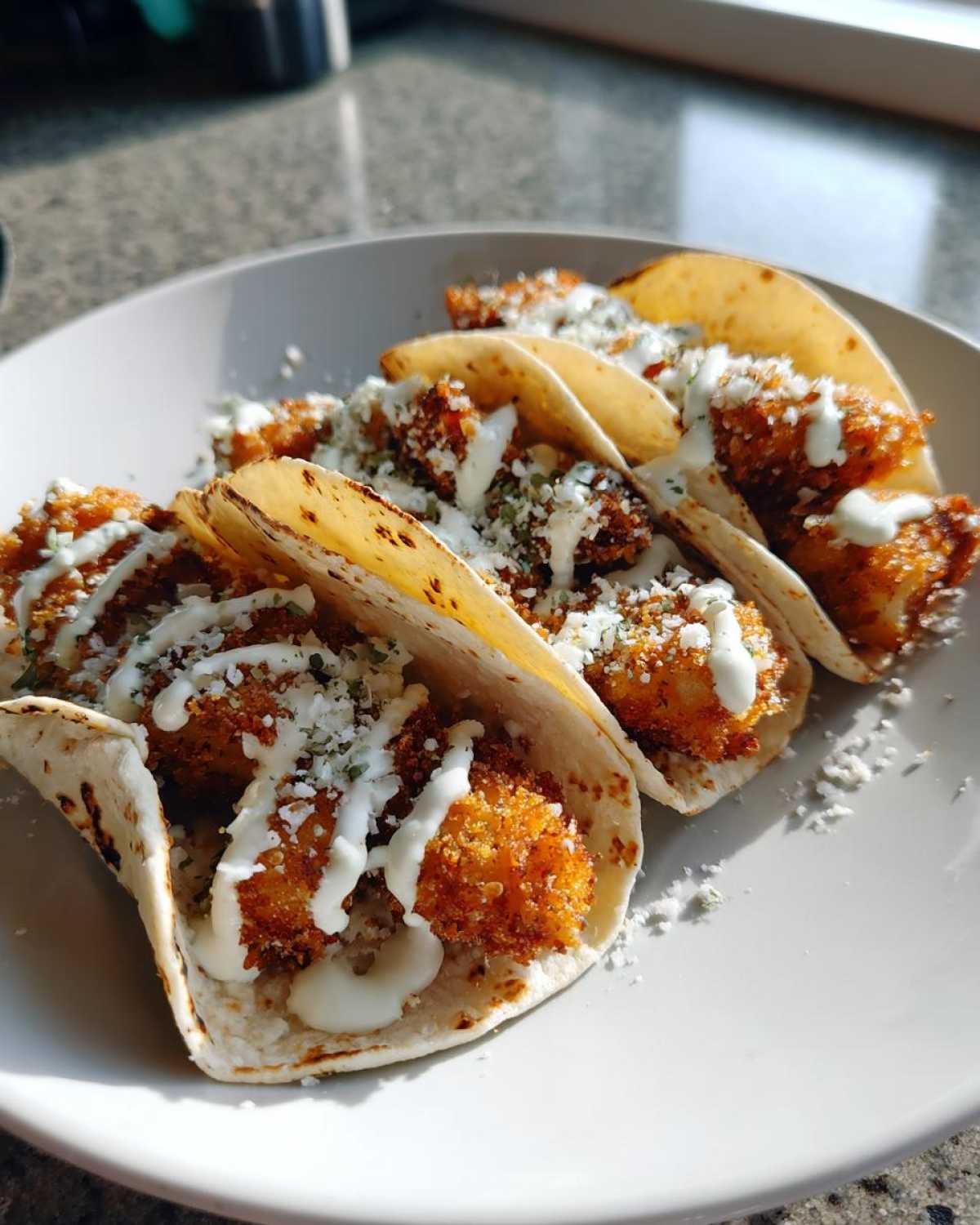 Four Crispy Chicken Tacos topped with white sauce drizzle and cotija cheese on a white plate.