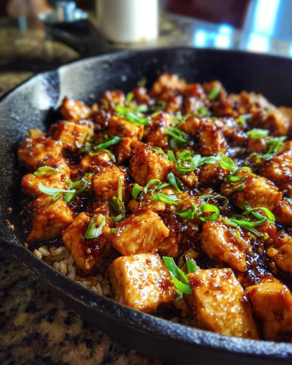Close-up of cubed chicken pieces coated in a rich glaze served over rice in a black skillet, topped with sliced green onions for Honey Garlic Chicken Skillet with Rice.