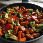 Close-up of glistening Honey Soy Chicken Skillet with Veggies including broccoli, carrots, and red peppers in a dark pan.
