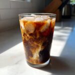 A tall glass of Iced Vanilla Chai Latte with ice cubes and creamy swirls of milk mixing into the dark chai concentrate.