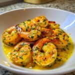 Close-up of cooked shrimp coated in a rich, yellow lemon butter sauce with visible garlic and parsley, served in a white bowl.