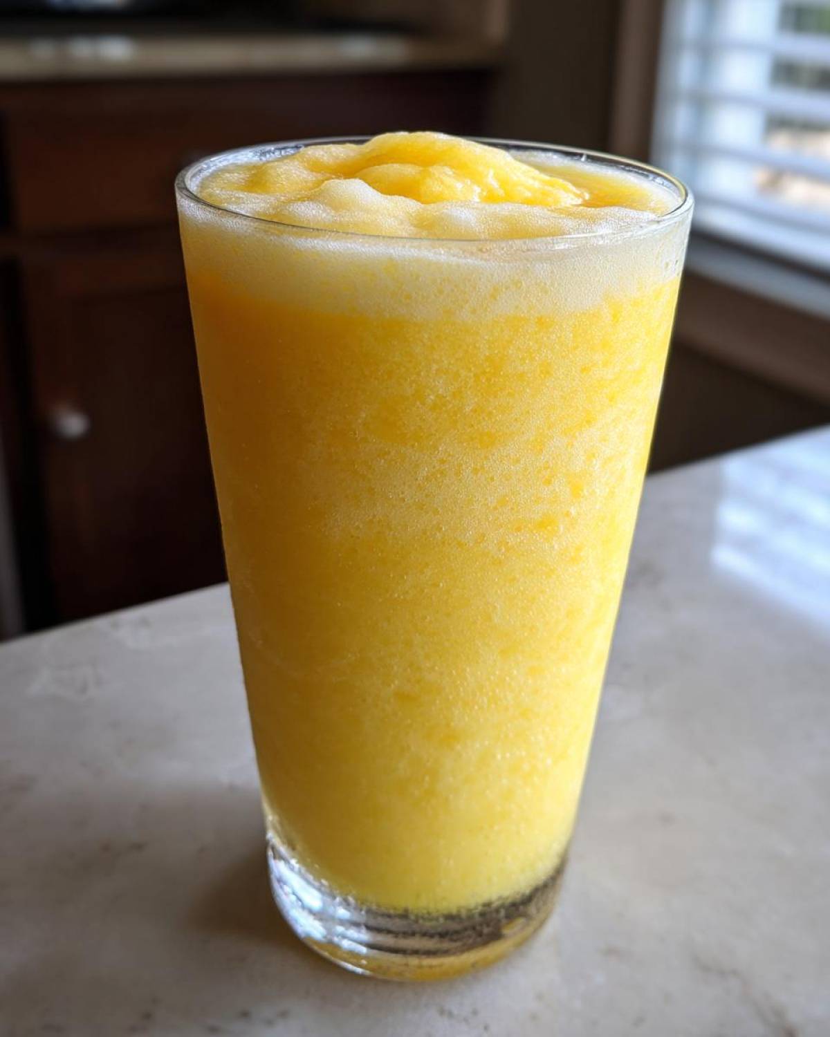 A tall glass filled with a thick, bright yellow Pineapple Coconut Smoothie, topped with foamy texture.